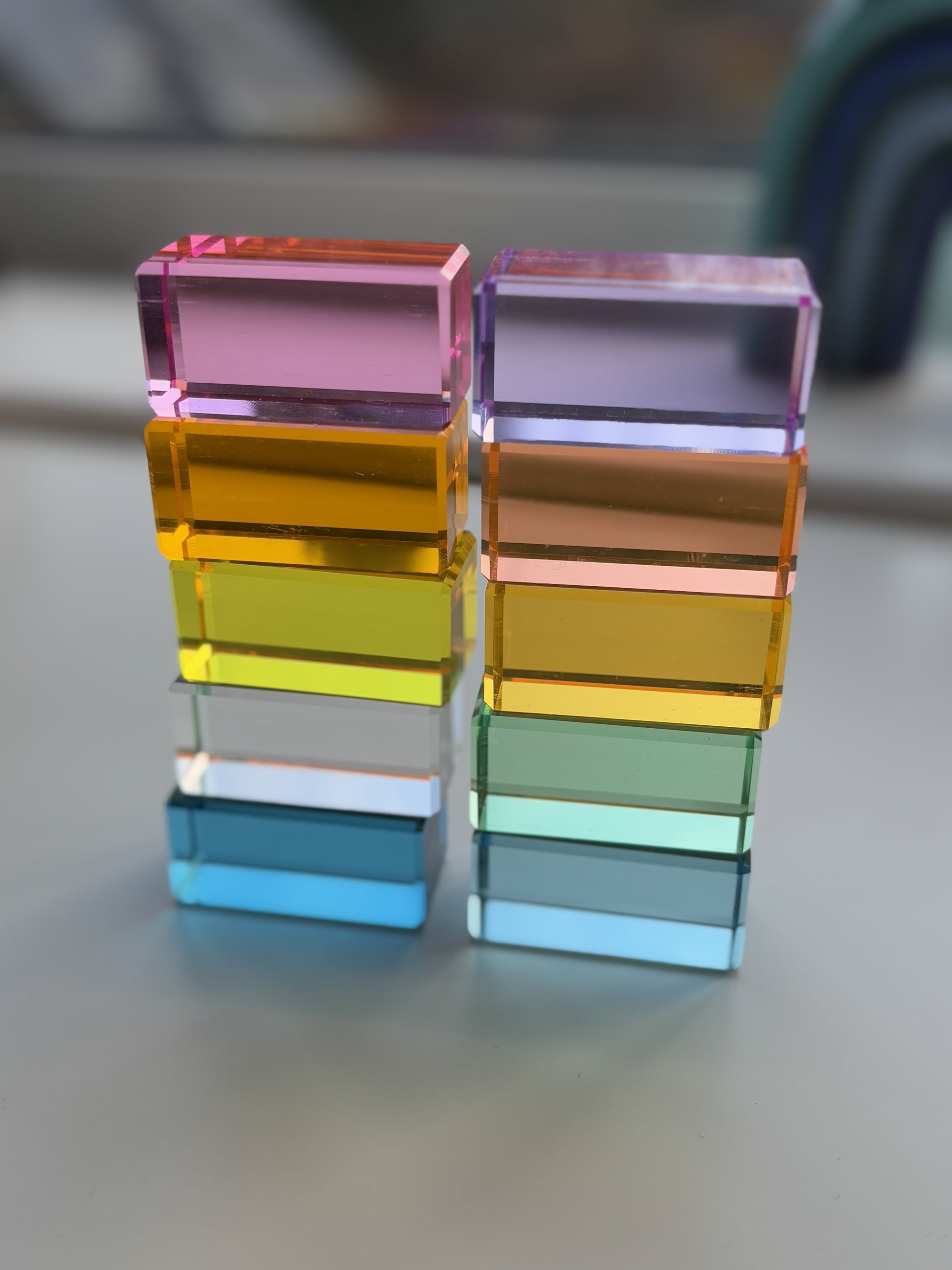 A set of 10 building blocks in rectangle shape stacked in two towers of 5 placed on a white surface. They are made from pastel coloured acrylic which refracts and reflects the spectrum of pastel colours on to the white surface.
