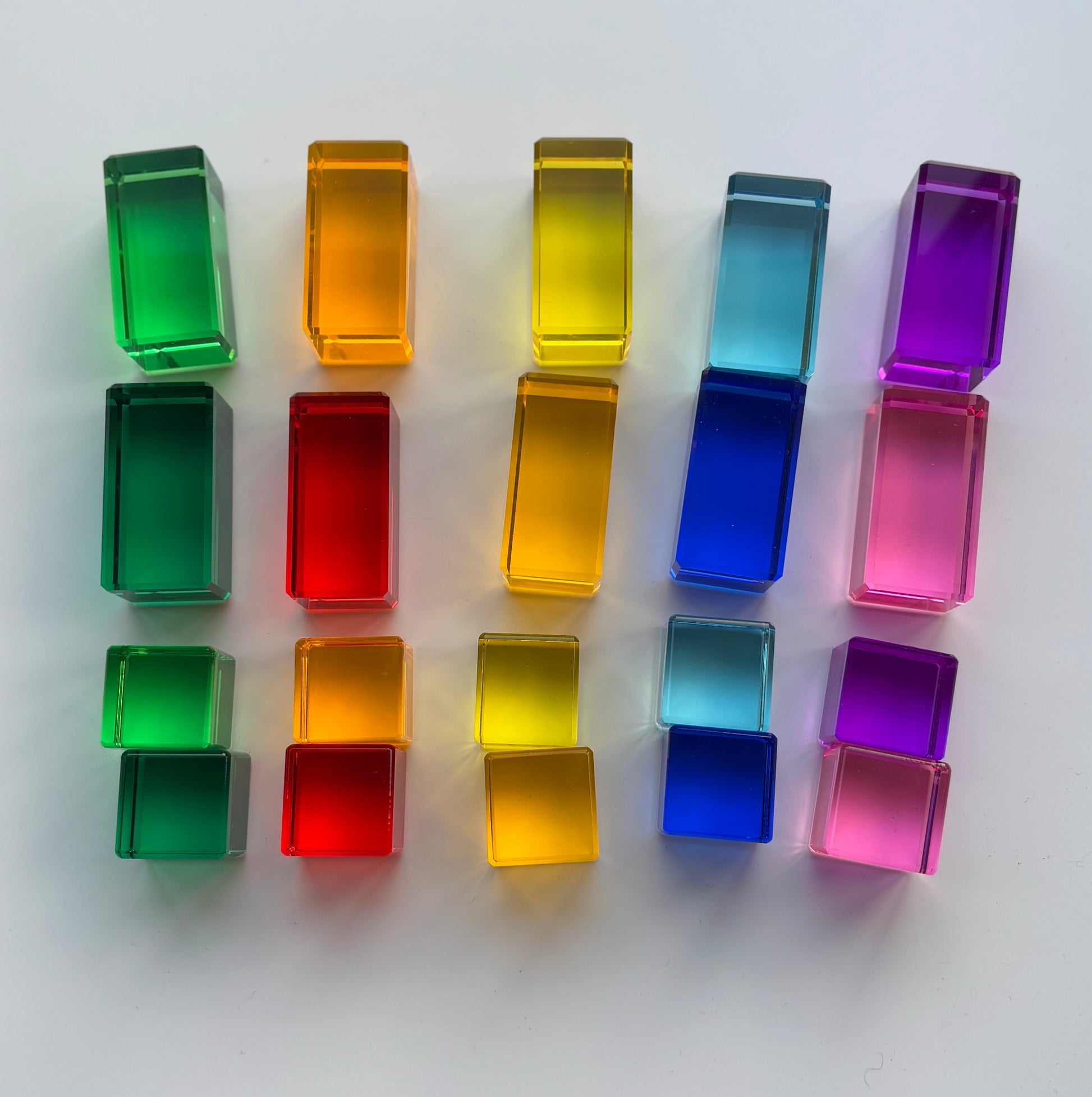 A set of Rainbow Cube and Rectangle Blocks arranged in a colour gradient in front of a window with the light penetrating through to display the colourful shadows. The acrylic material reflects the light. Infinite Wonder Acrylic Building Blocks - a great addition to any play space to build children's creativity and skill building.