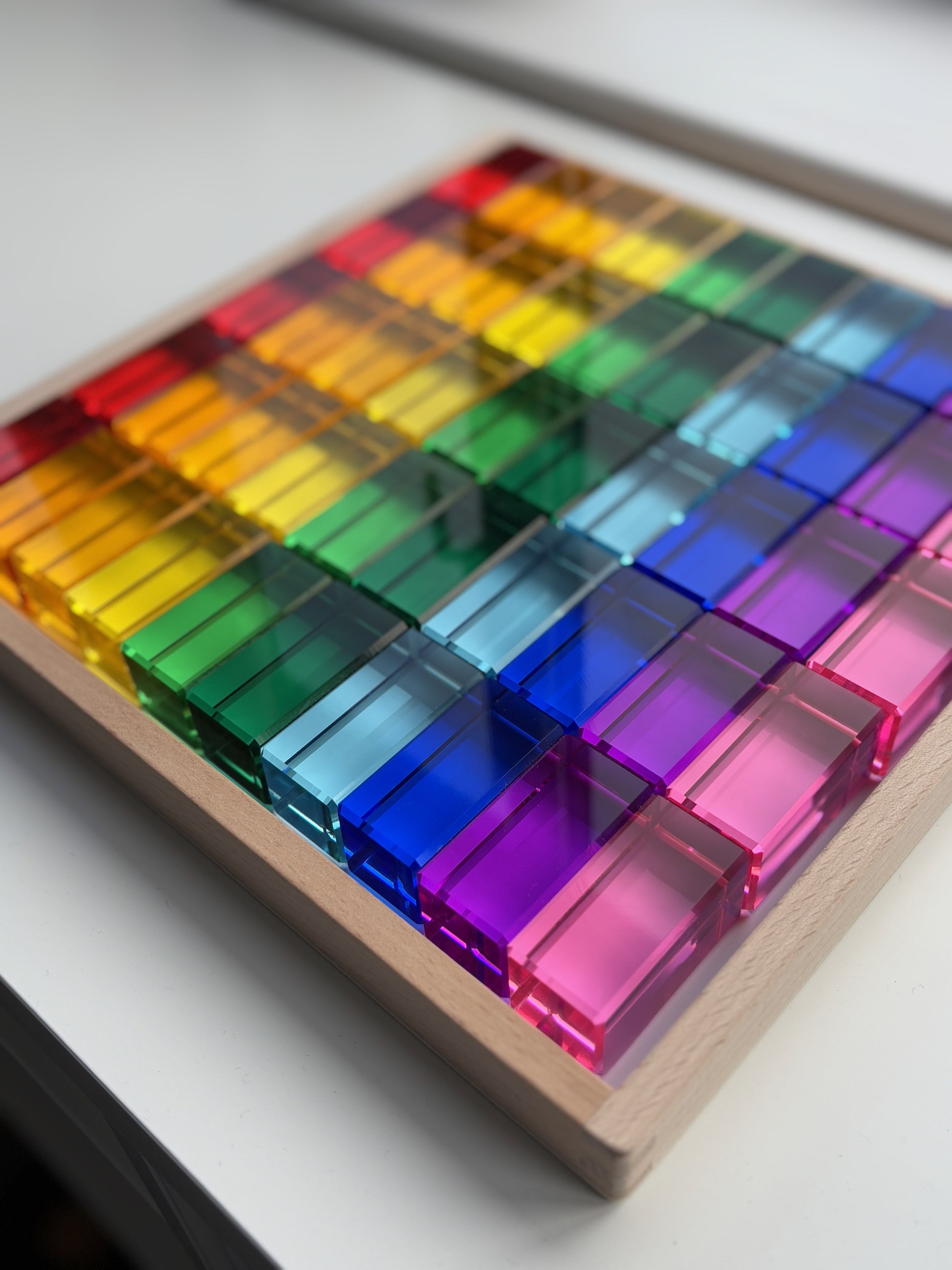 A set of 100 Rainbow Rectangle Blocks arranged in a colour gradient in a wooden frame tray. The acrylic material reflects the light. Infinite Wonder Acrylic Building Blocks - a great addition to any play space to build children's creativity and skill building.
