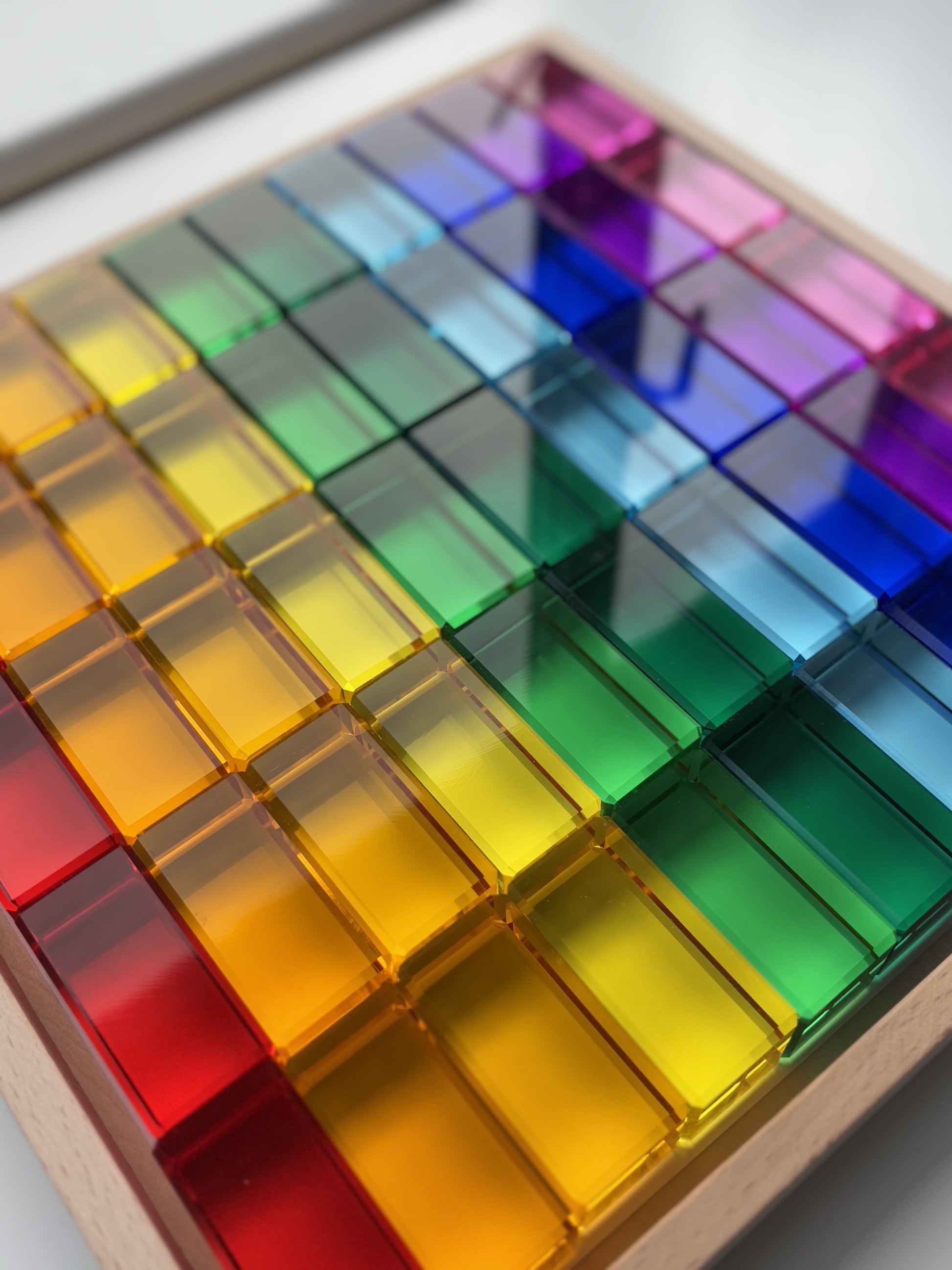 A photo of the Infinite Wonder Rectangle Rainbow Building Blocks in a wooden rimmed tray. The photo showcases how beautiful the smooth, translucent surface of each block captures and reflects the light.