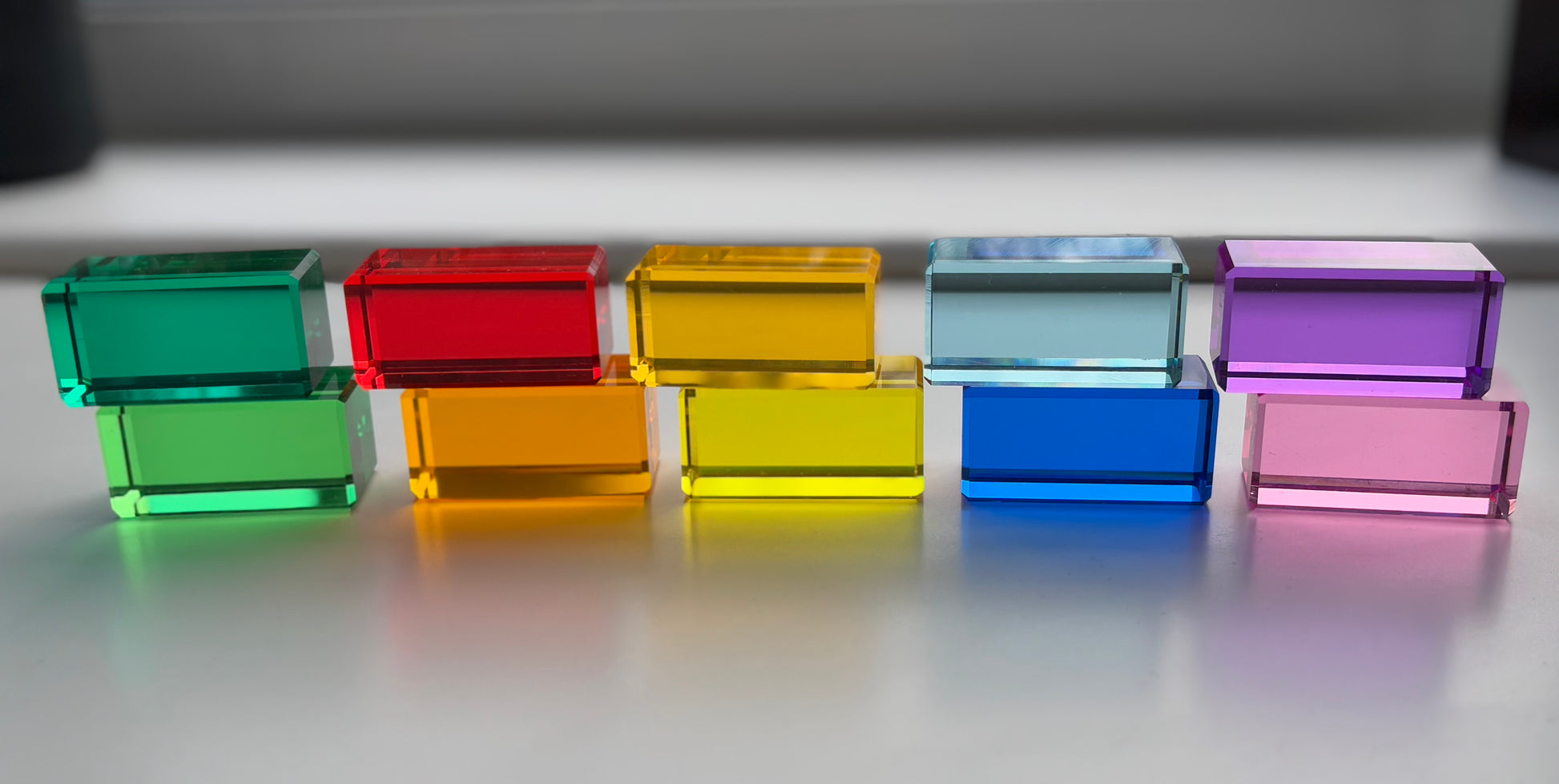 A set of Rainbow Rectangle Blocks arranged in a colour gradient in front of a window with the light penetrating through to display the colourful shadows. The acrylic material reflects the light. Infinite Wonder Acrylic Building Blocks - a great addition to any play space to build children's creativity and skill building.