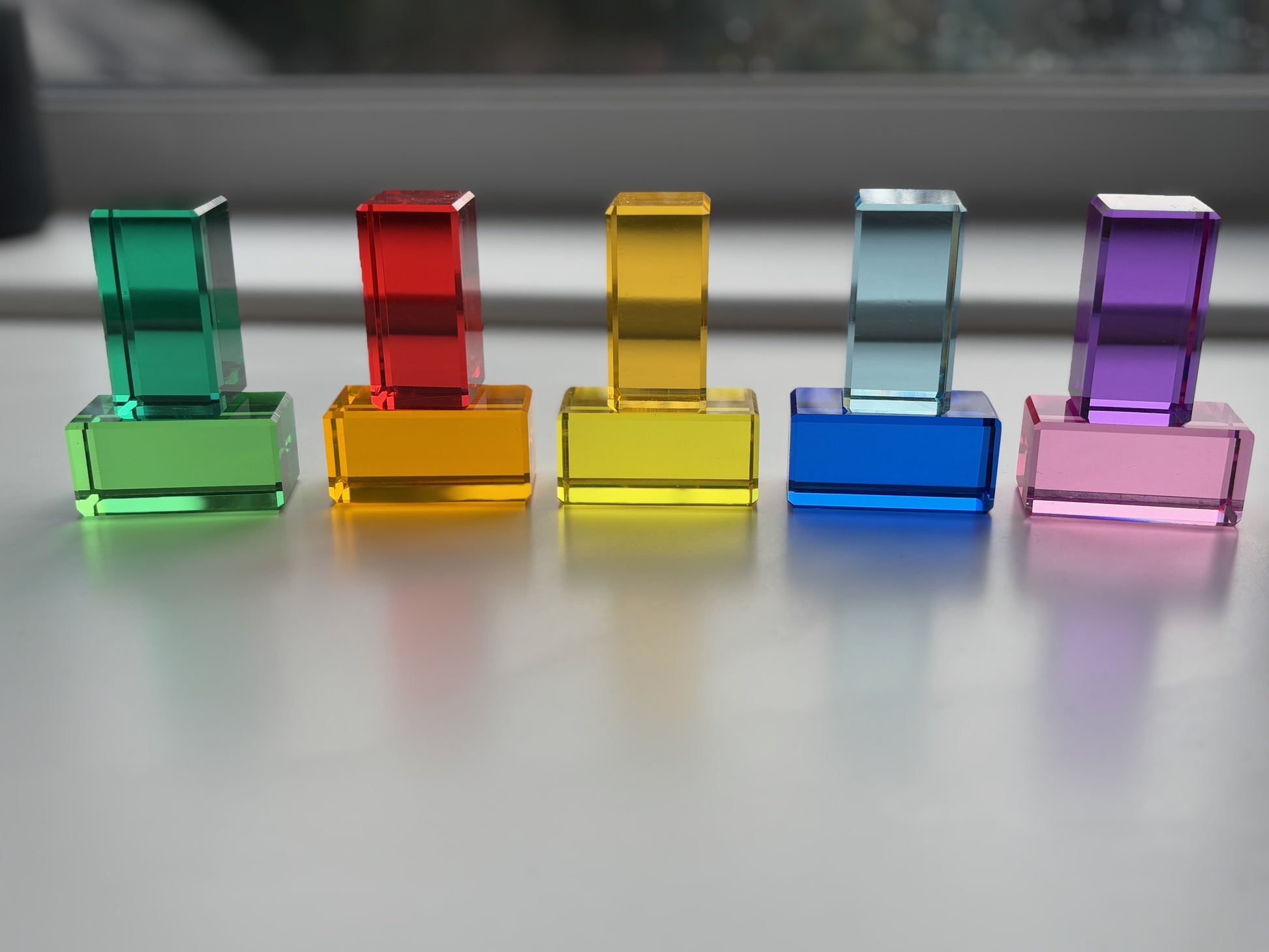 A set of Rainbow Rectangle Blocks arranged in a colour gradient in front of a window with the light penetrating through to display the colourful shadows. The acrylic material reflects the light. Infinite Wonder Acrylic Building Blocks - a great addition to any play space to build children's creativity and skill building.