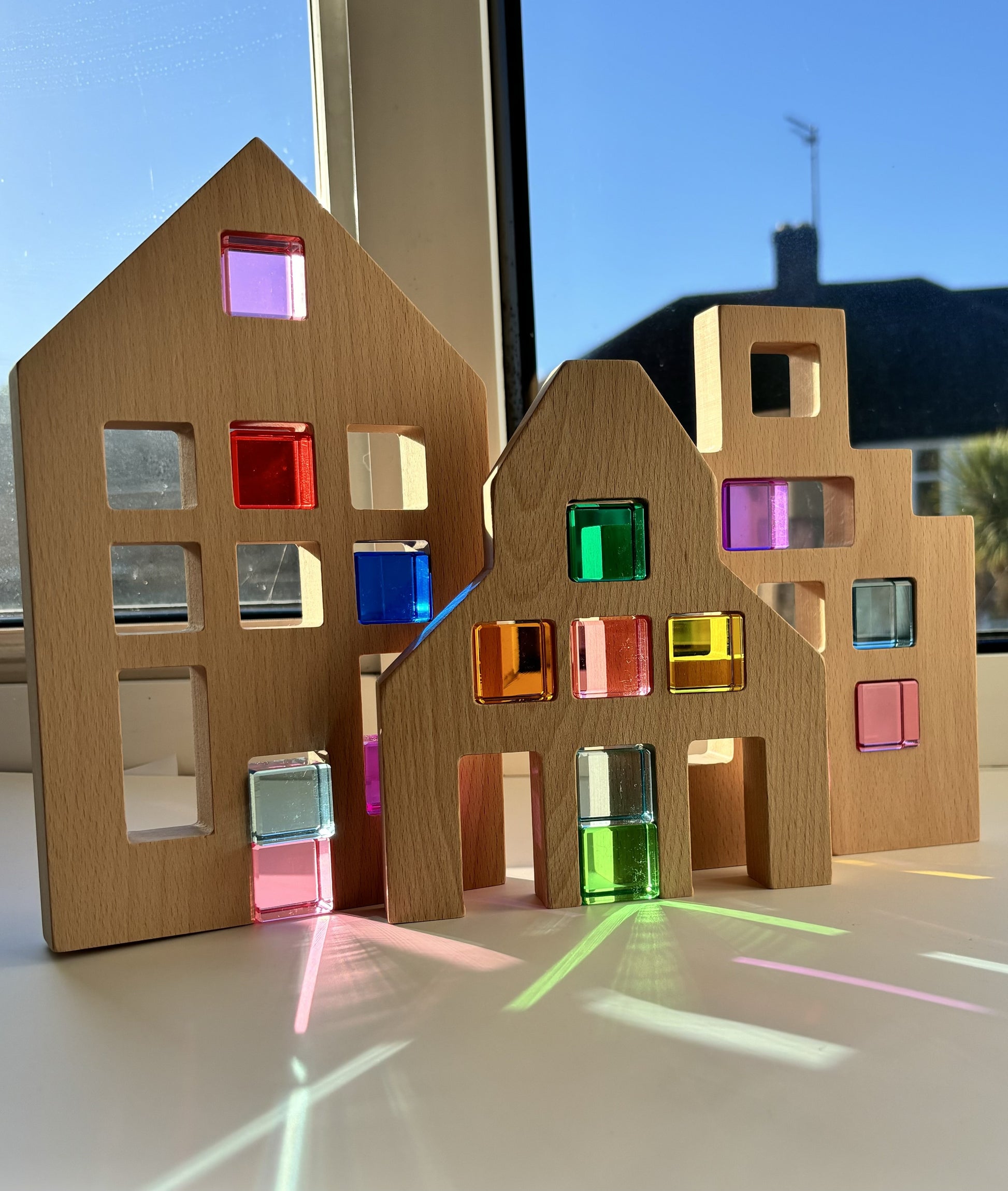 3 Wooden Window Houses sit on a window ledge with a number of rainbow acrylic blocks placed in each window. The rainbow colours of each block are reflected and refracted on to the window ledge underneath.
