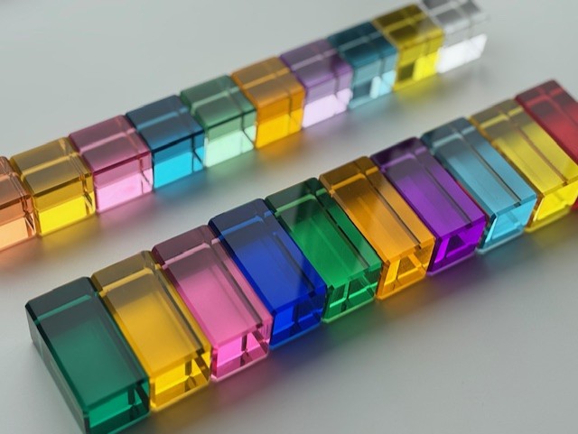 Pastel and rainbow transparent acrylic blocks arranged in a pattern on a white surface, each colour reflects through the block on to the white surface.