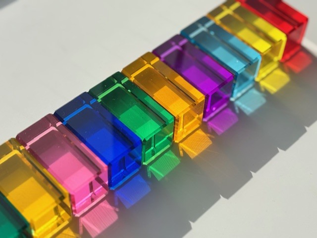 Rainbow transparent acrylic blocks arranged in a pattern on a white surface, each colour reflects through the block on to the white surface.